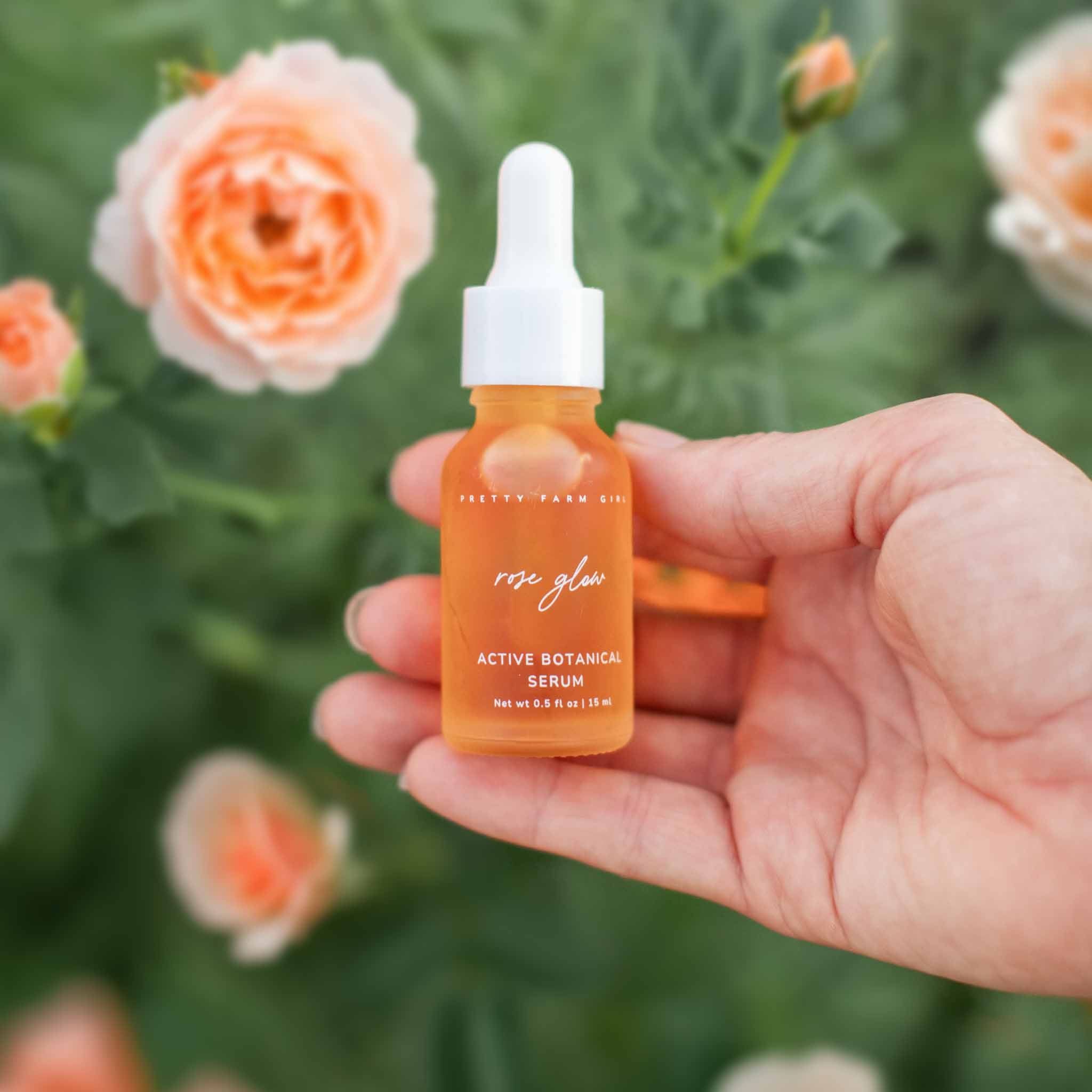 Hand holding Rose Glow Active Botanical Serum bottle with white dropper against blooming peach roses in garden background